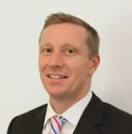 Smiling man in a business suit with a striped tie against a plain background.