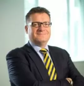 Man in suit with a yellow striped tie and glasses, smiling and standing with arms crossed.