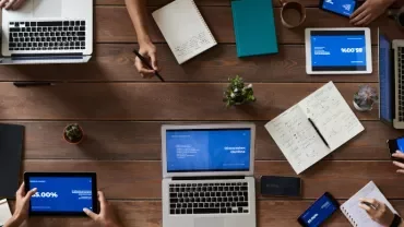 Top view of people working on laptops and tablets at a wooden table.