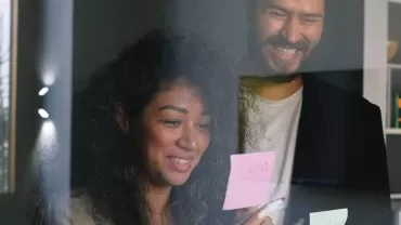 Two people smiling and reading sticky notes on a glass wall in an office.