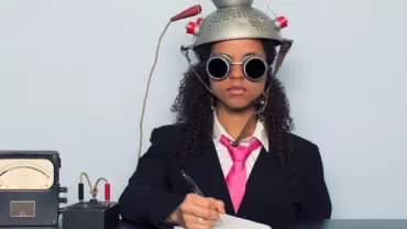 Person wearing a suit, pink tie, and goggles, with a strainer helmet connected to a device, writing in a notebook.
