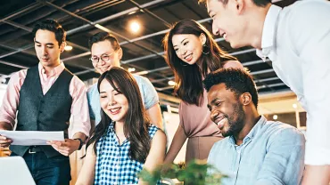 A group of colleagues collaborating at a desk, smiling and looking at a laptop.