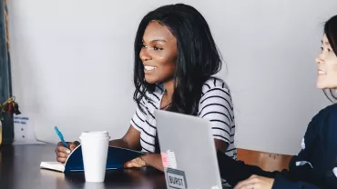 Two women at a table with laptops and coffee, engaged in discussion.
