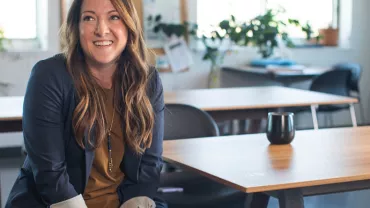 Smiling person sitting in a modern office with wooden tables and plants.