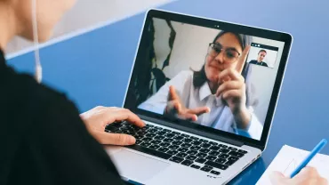 Person on laptop video call taking notes with a pen.