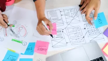 People brainstorming at a desk with wireframe sketches and colorful sticky notes.