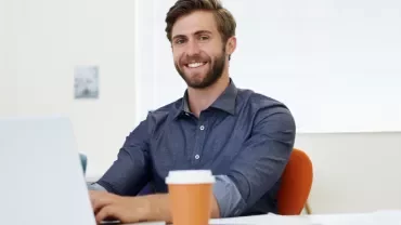 Smiling man in a blue shirt working on a laptop at a desk with a coffee cup.