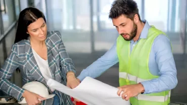 Two professionals reviewing building plans in an office corridor.