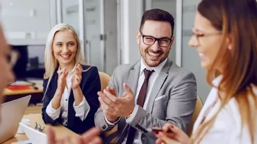 Group of professionals smiling and clapping in a modern office meeting.