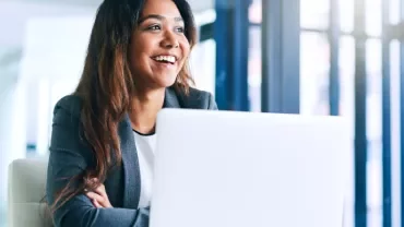 Woman in a suit smiling while working on a laptop in a bright office.