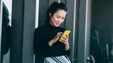 Woman smiling while using a yellow smartphone outside a building.