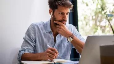 Man wearing earphones working on a laptop while taking notes.
