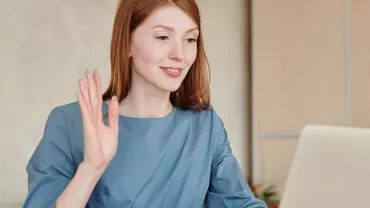 Woman in blue shirt waving at laptop during video call.