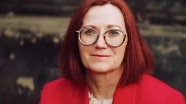 Woman with red hair and glasses wearing a red jacket.