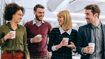 Four colleagues walking and holding coffee cups while smiling.