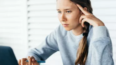 Young woman focuses intently on laptop screen, holding pen.