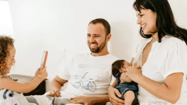 A family sits together on a bed; the girl uses a smartphone.