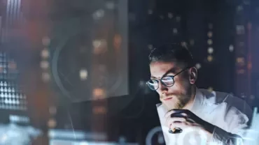 Man working on laptop, analyzing data on screen in dark room with graphs.