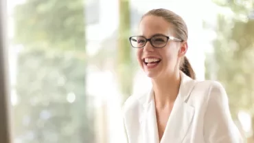 Woman in glasses laughing while sitting in a bright room.