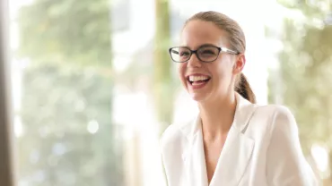 Woman in glasses laughing while sitting in a bright room.