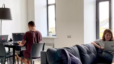 Three people working on laptops in a bright room with large windows.