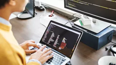 Person coding on a laptop with a larger screen displaying code.