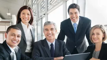 A diverse group of business professionals smiling around a laptop in a modern office.