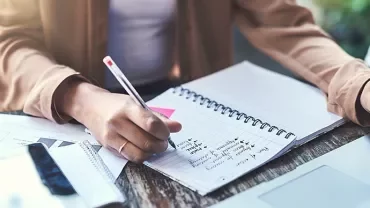 Person writing notes in a spiral notebook at a desk with documents and a laptop.