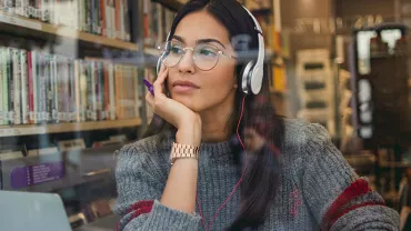 Woman wearing headphones looking out a library window, with books in the background.