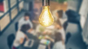 Close-up of a lit light bulb with blurred people working in the background.