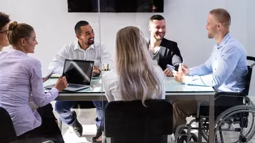 A diverse group of colleagues smiling in a meeting around a glass table.