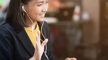 Woman smiling and waving during a video call, wearing earphones, with a blurred background.