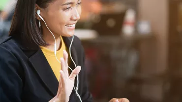 Woman smiling and waving during a video call, wearing earphones, with a blurred background.