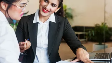 Two women having a discussion at a desk in an office setting.