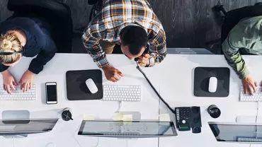 Overhead view of three people working at office desks with computers, using headsets and keyboards.