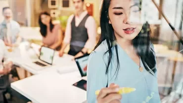 Woman writing with marker on glass, with colleagues in conference room.