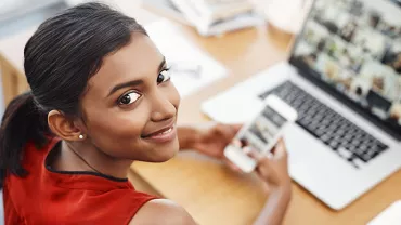 Woman smiling while holding phone at desk with laptop open.