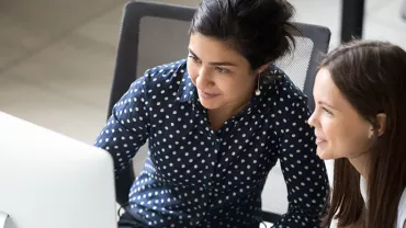 Two women smiling and working together at a computer desk in an office setting.
