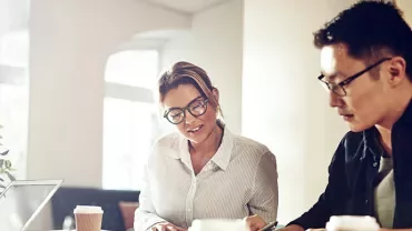 Two people working together on documents at a table with coffee and a laptop.