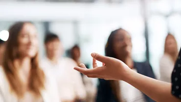 A speaker gesturing in front of a blurred audience.