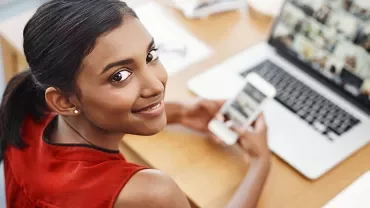 Woman in red top smiling, holding a phone, looking at a laptop with images.