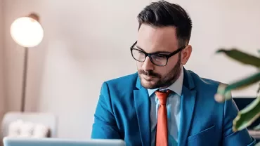 Man in blue suit and glasses working on a laptop in a modern office.