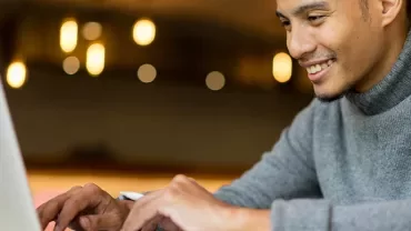 Man in gray sweater smiling while using a laptop.