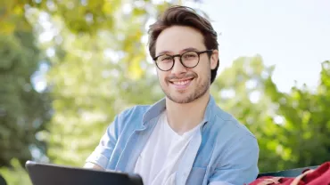 Smiling person with glasses sitting outdoors, holding a tablet.