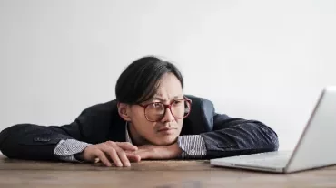 Man with glasses looking at a laptop on a wooden table.