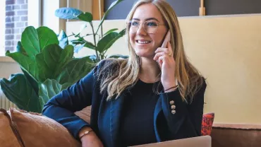 Smiling woman in glasses talking on the phone, sitting on a couch with a laptop nearby.