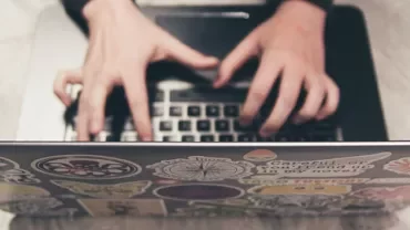 Hands quickly typing on a laptop covered with various colorful stickers.