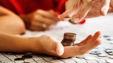 Hand holding a stack of coins, with another hand adding a coin to the stack on a table.