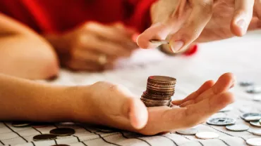 Hand holding a stack of coins, with another hand adding a coin to the stack on a table.