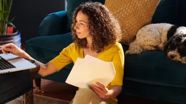 Woman working on a laptop at home with a dog sleeping on the couch.